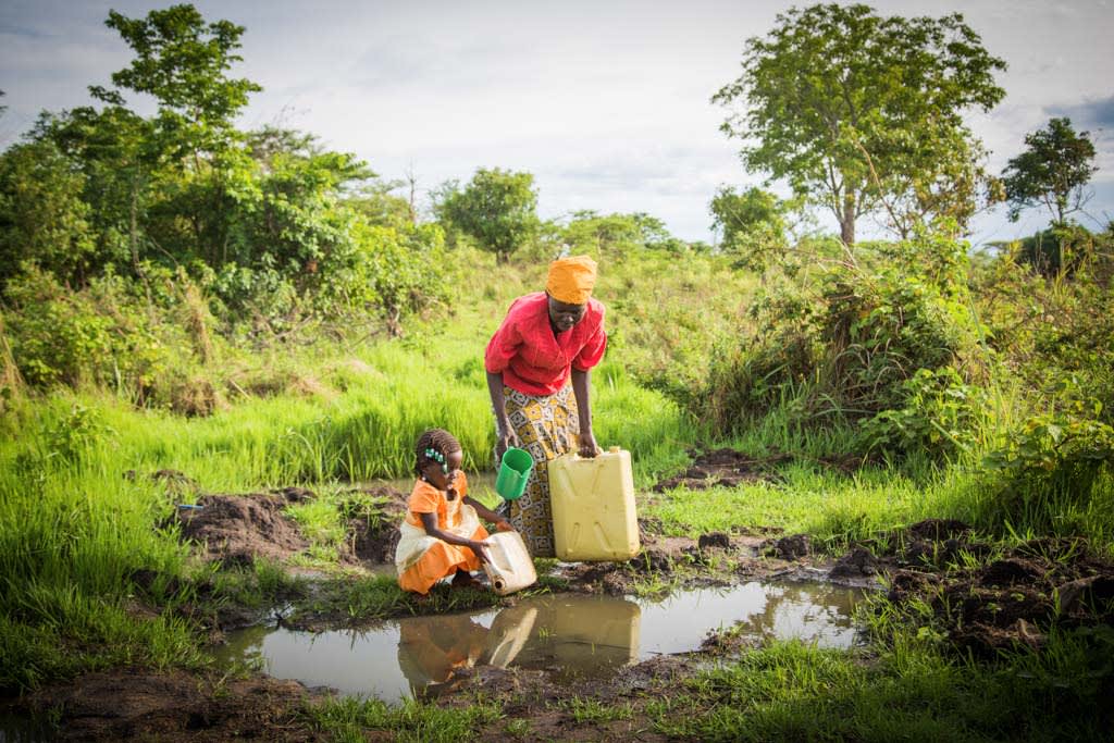 Betty, a girl, child, wearing an orange and white dress, bends over to fetch water with a plastic container beside her mother as they are getting water in plastic buckets and containers, daily household chores, parent, adult woman, Evelyn, who is wearing a yellow scarf, turban, head wrap on her head, a red shirt and long pattern skirt, together at a small water source, large puddle, large mud puddle of collected rain water on the ground, with the mother using a green cup in her hands to retrieve the water to pour into the containers. This appears to be an unsanitary water source, potentially unhealthy, unsafe, and not clean drinking water in a large puddle on the ground. They are surrounded by trees in the background, and dark mud on the ground and grass.
